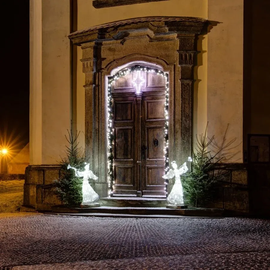 Deux anges LED blanc glacé à la trompette devant une porte baroque ornée de petits sapins de Noël, vue de nuit de l'entrée